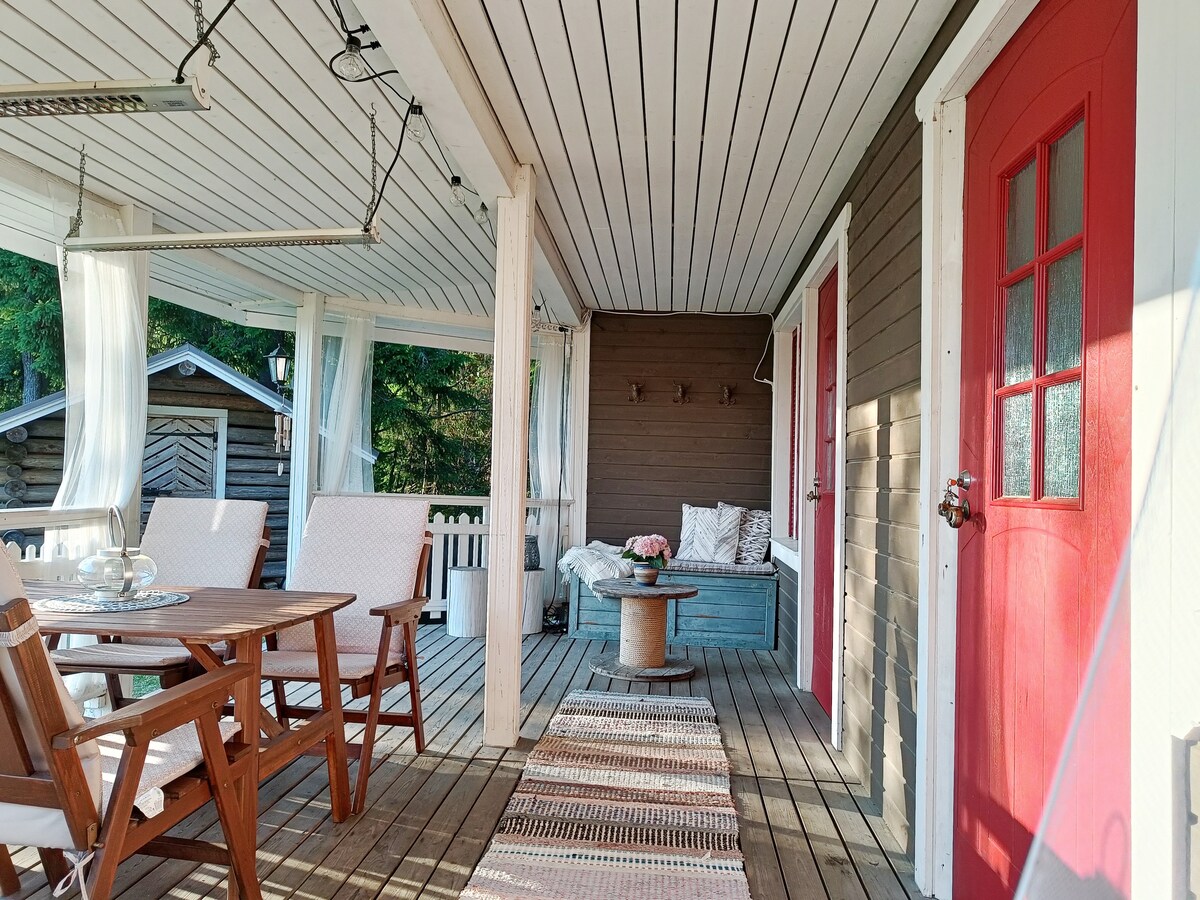 A covered porch area is visible, featuring a wooden dining table with four chairs and a decorative centerpiece. Two red doors lead into the interior space, which includes a cozy seating area with a sofa and soft cushions. A patterned rug adds warmth to the wooden floor.