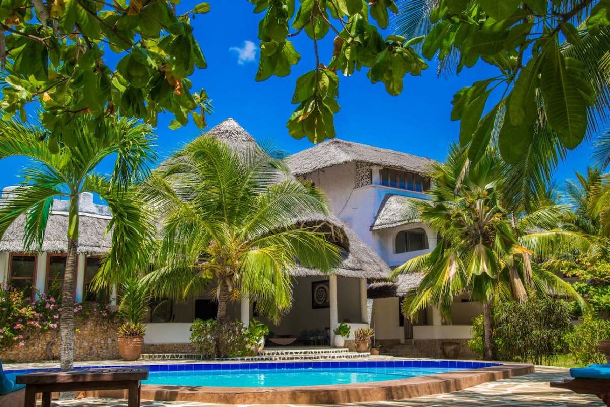 A serene villa is set against a clear blue sky, showcasing a thatched roof and surrounded by lush palm trees. A private swimming pool sits in the foreground, framed by greenery, creating a tranquil outdoor retreat.