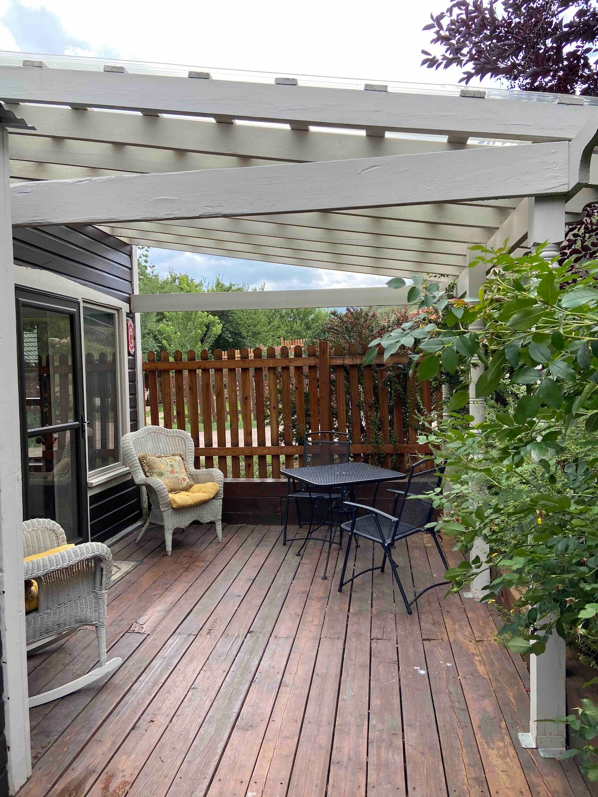 An outdoor patio area features a wooden deck surrounded by greenery. Two comfortable chairs are positioned beside a small round table. A pergola overhead provides shade, and a fenced yard is visible in the background.