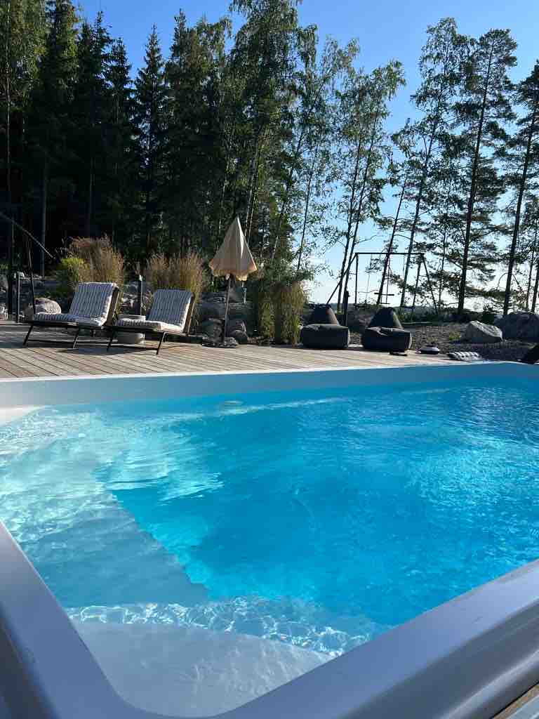 A warm, inviting heated pool is surrounded by a spacious deck, complemented by lounge chairs. Verdant trees and a clear blue sky provide a serene backdrop, while natural vegetation enhances the relaxing atmosphere of the outdoor space.