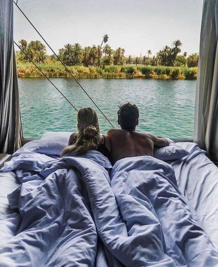A serene view is captured from a spacious bed with light blue bedding, facing the river. The tranquil water reflects greenery and palm trees along the banks, creating a calming ambiance. Two individuals are seen enjoying the peaceful scenery together.