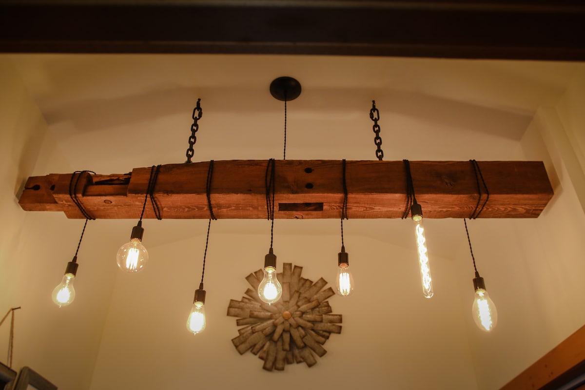 A rustic wooden light fixture hangs from the ceiling, featuring a long beam with attached pendant bulbs. The warm glow of the bulbs contrasts against the light-colored wall, while a decorative wall piece is visible below, enhancing the cozy ambiance of the space.