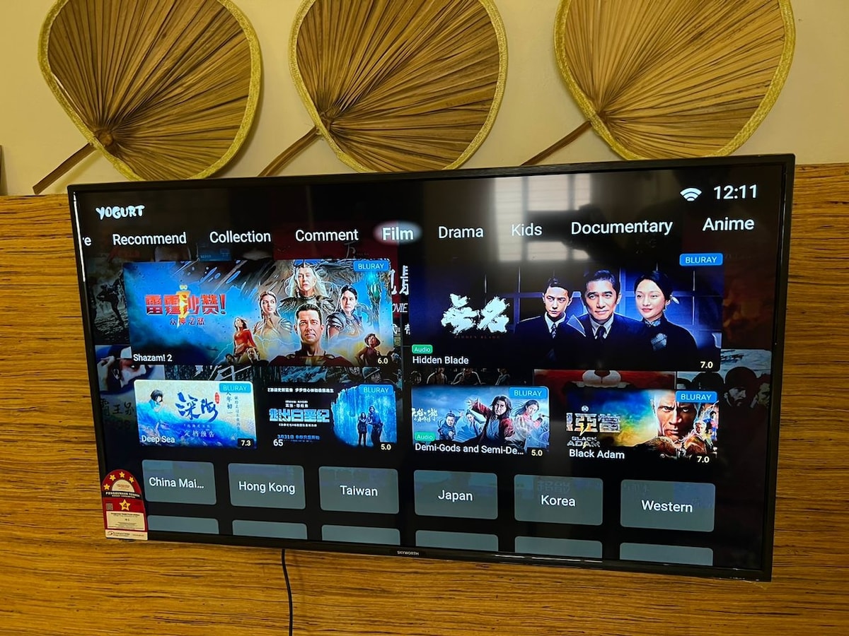A large flat-screen TV is mounted on a wooden wall, featuring a user interface that organizes content into categories such as Film, Drama, Kids, and Documentary. Bold images from various movies and shows are displayed, with a warm background created by decorative wall elements.