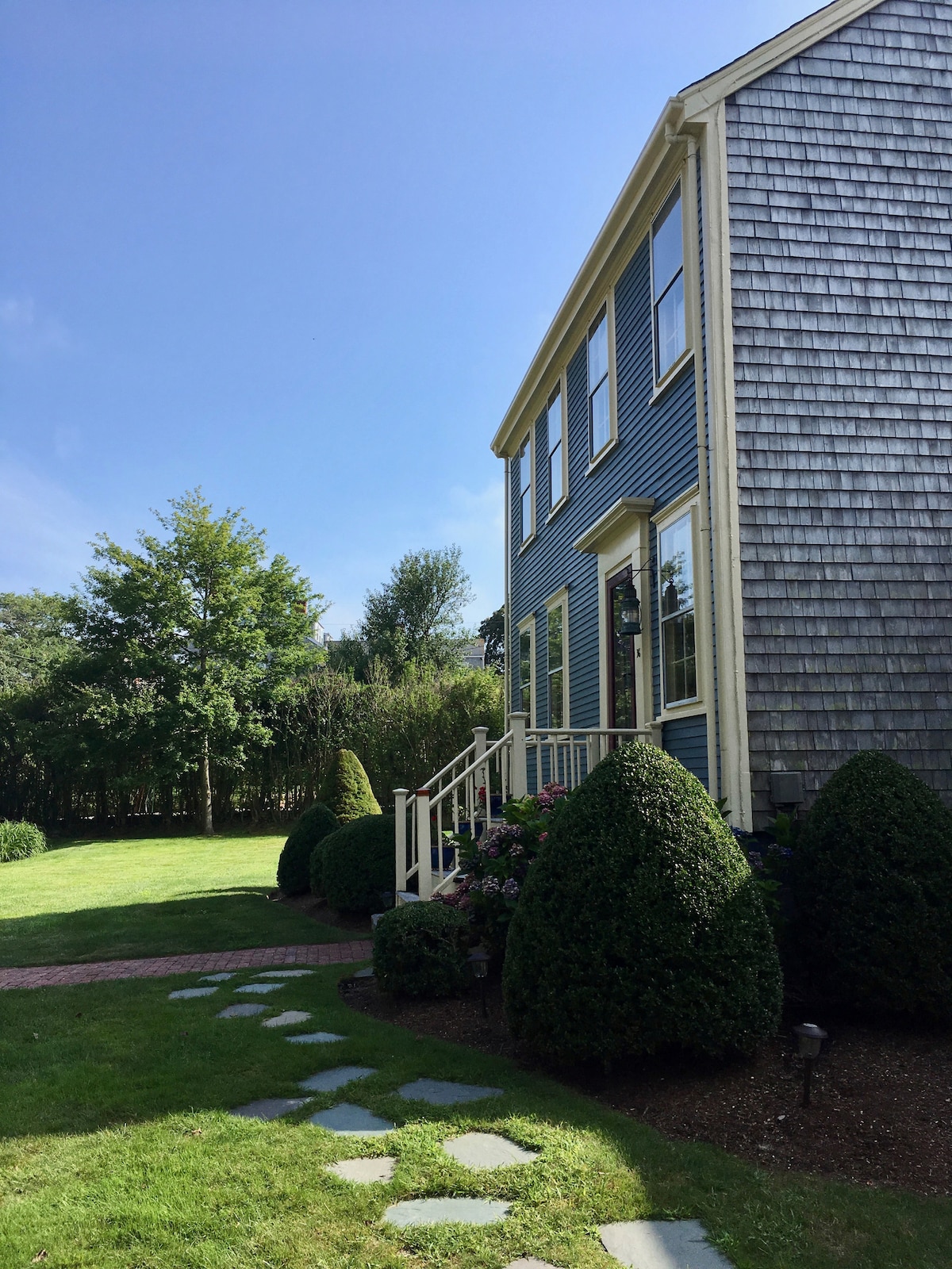The exterior of the home is shown, highlighting a charming blue facade and a well-maintained entrance. A stone path leads to the porch, surrounded by neatly trimmed shrubs and vibrant greenery in the yard, all under a clear blue sky.