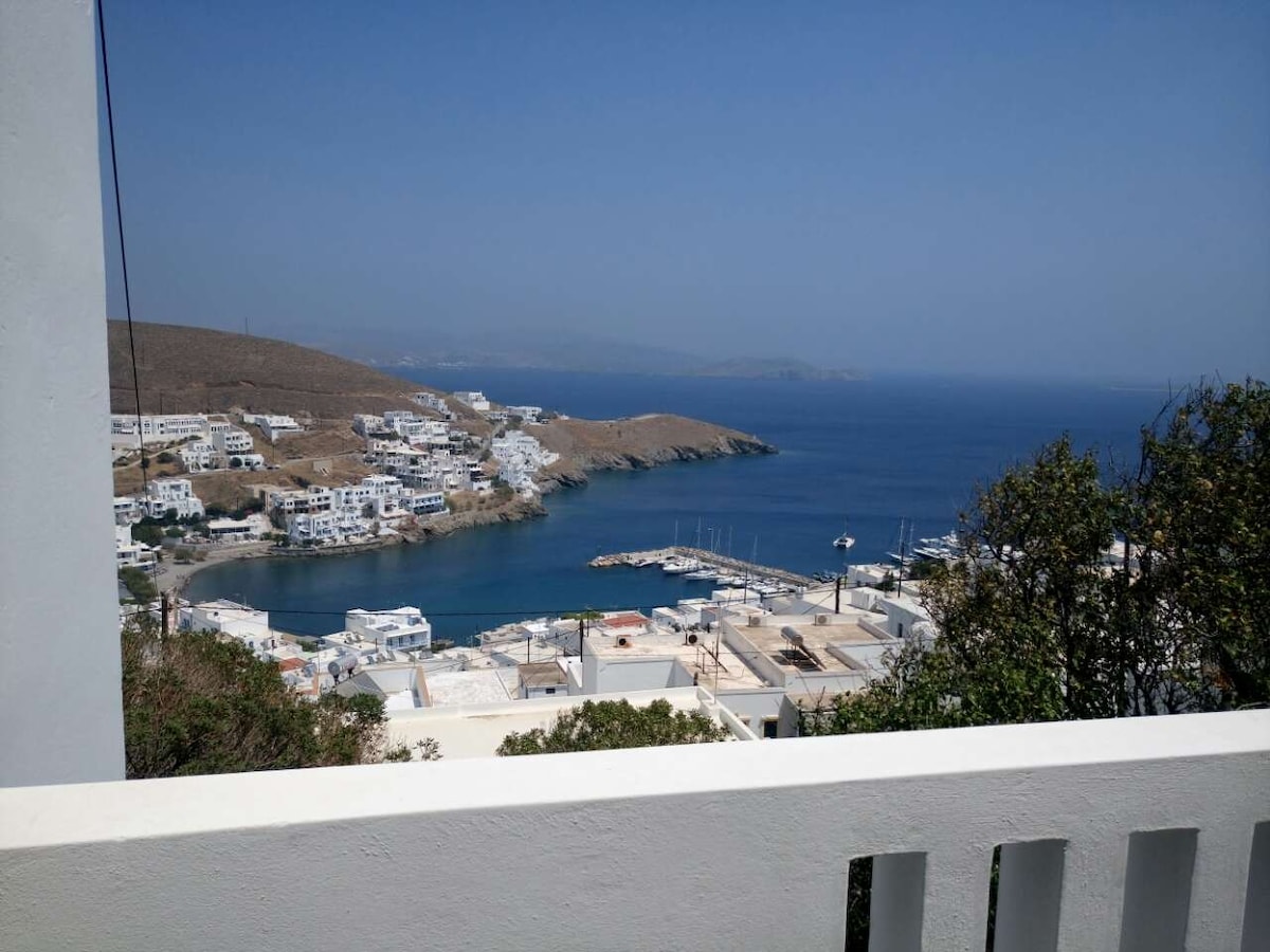 A scenic view of a coastal village is displayed, featuring whitewashed buildings nestled along the shore. The clear blue sea contrasts with the surrounding hills, while boats are docked in the harbor, illustrating a serene coastal landscape.