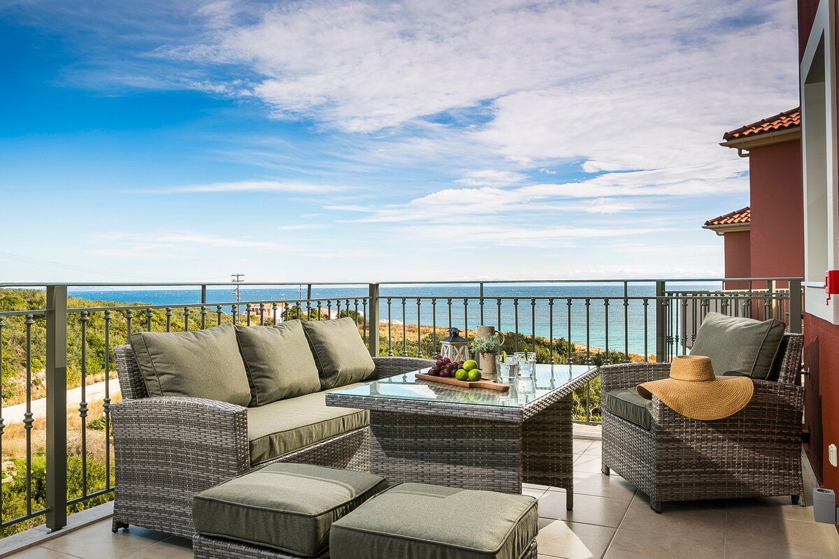 The veranda showcases comfortable seating with plush cushions, a glass-topped table adorned with refreshments, and a wide view of the Ionian Sea. The outdoor area is accented by a sun hat, suggesting a relaxing atmosphere to enjoy the surrounding natural beauty.