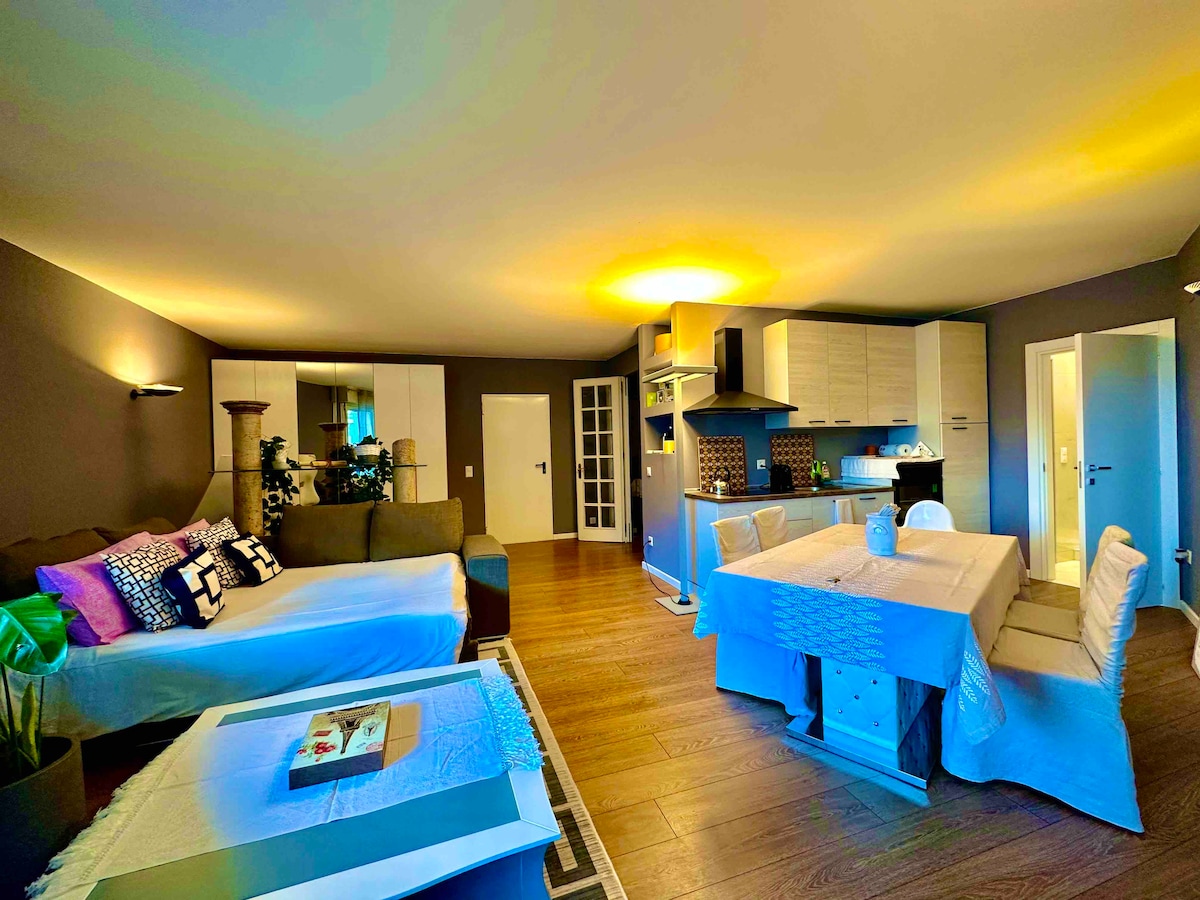 An open-plan living area features a seating arrangement with a light-colored sofa and decorative cushions. A dining table with textured tablecloth is set for meals. The kitchen area showcases modern cabinetry. Warm lighting enhances the spaciousness, and light wooden flooring complements the neutral wall tones.