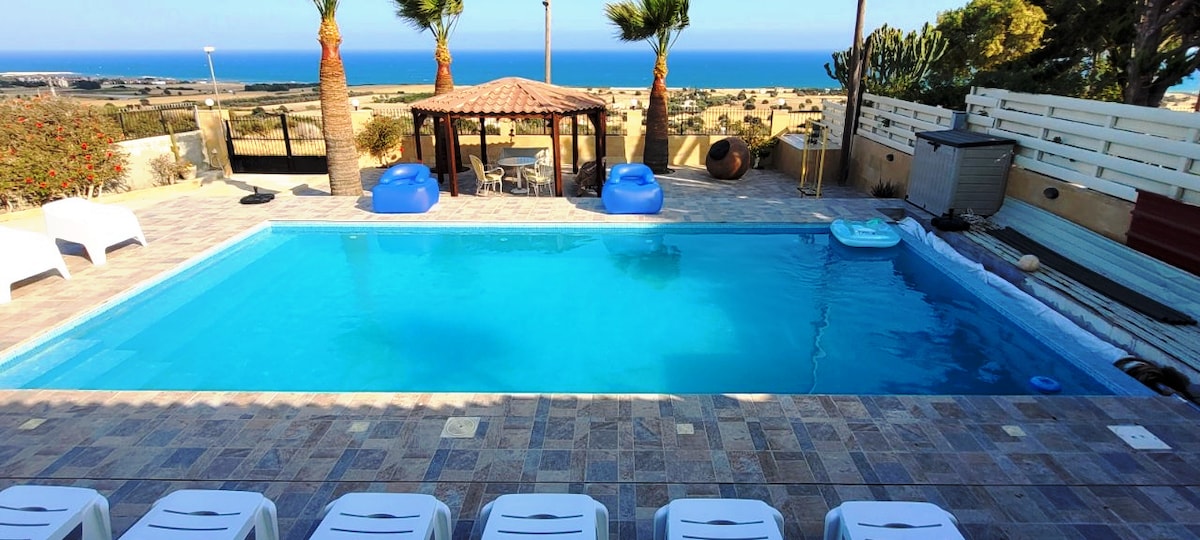 A tranquil outdoor area features a swimming pool bordered by lounge chairs. A gazebo with palm trees stands nearby, offering shade. The expansive sea view is visible in the background, blending with the blue sky and horizon.