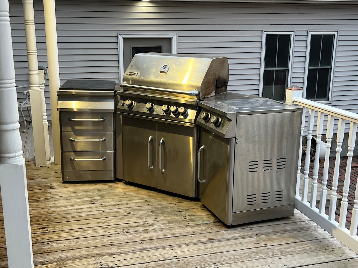 A stainless steel grill is prominently placed on a wooden deck, accompanied by a matching cabinet. The grill features multiple burners and a large cooking surface, ready for outdoor meals. The deck's railing and wooden planks provide a comfortable space for gatherings.
