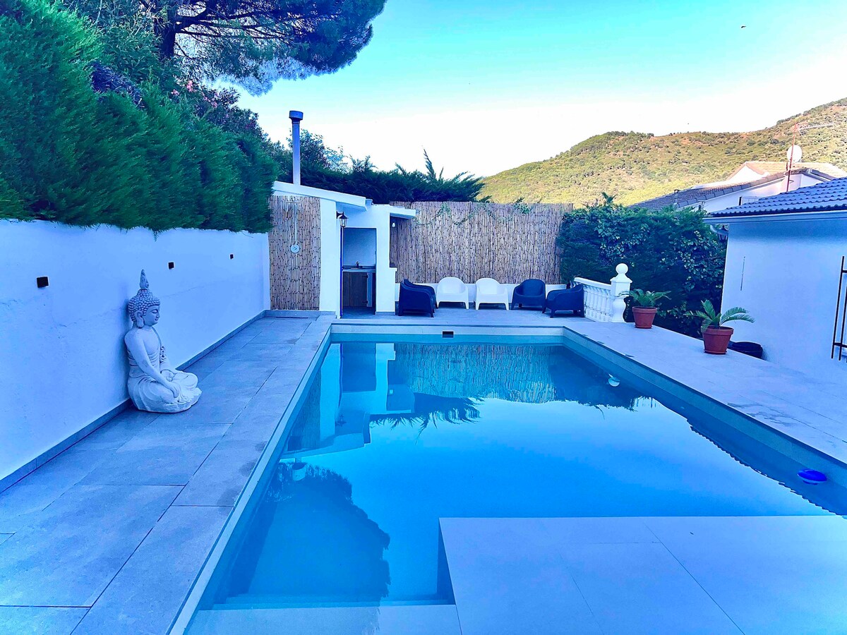 A private pool area features clear blue water, surrounded by elegant stone tiles. Potted plants add greenery, while a seated area is complemented by a soothing buddha statue. Fenced landscaping provides privacy against the backdrop of serene hills.