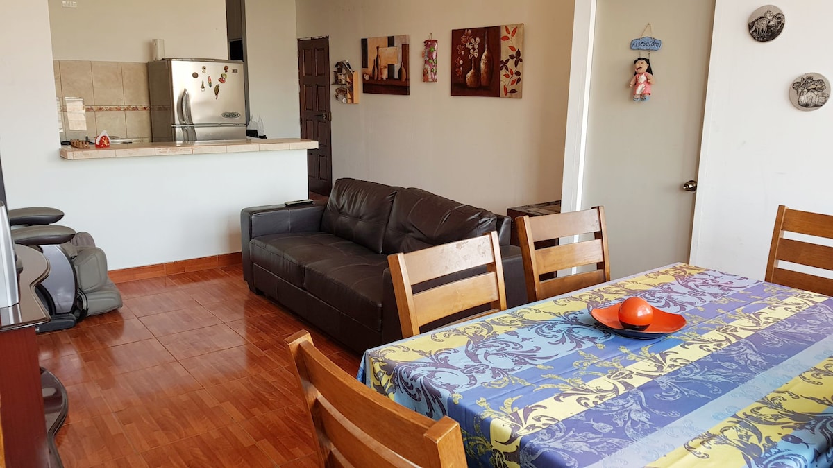 A comfortable living space featuring a dark leather sofa and a vibrant, patterned dining table with wooden chairs. The kitchen area is visible in the background, highlighting stainless steel appliances and light-colored countertops. Wall art adds a personal touch to the neutral-toned walls.