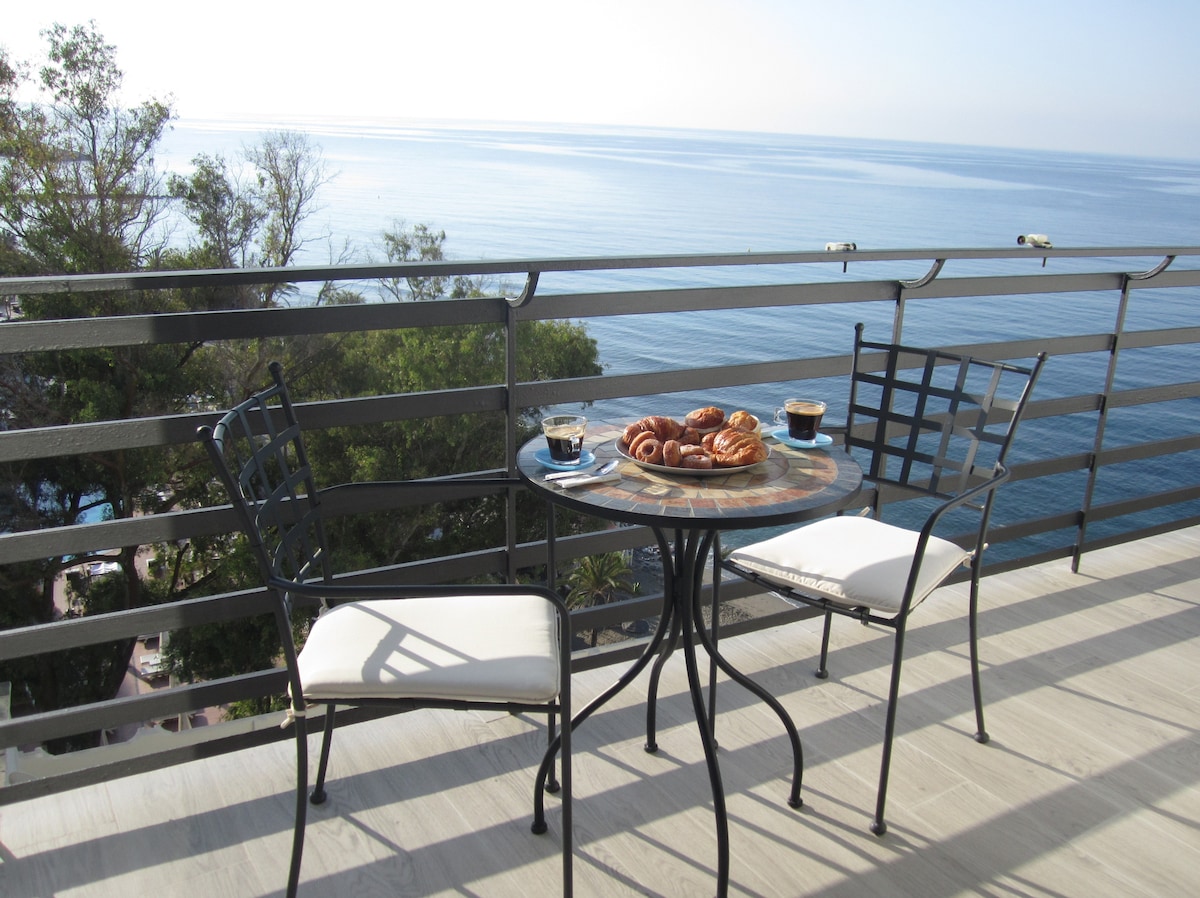 A balcony table with two chairs overlooks the sea. Fresh pastries and cups of coffee are arranged on the table. Lush greenery is visible in the background, complementing the expansive ocean view under a clear sky.