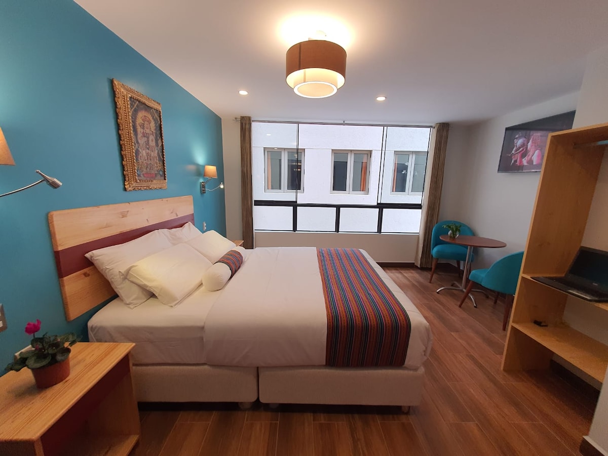 A bright hotel room features a spacious bed with a colorful throw and decorative pillows. A small table with a chair is positioned near a large window, allowing natural light to fill the space. An illuminated wall sconce adds to the cozy ambiance.