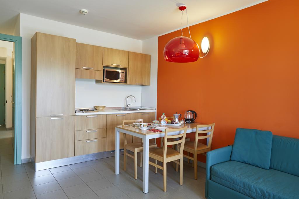 A compact kitchen area features modern wood cabinetry and a built-in microwave, complemented by an orange accent wall. A simple dining table set for four is present, alongside a blue sofa. Natural light enhances the inviting layout of the space.