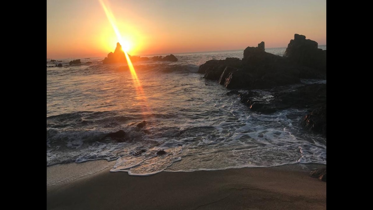 The image captures a serene beach scene at sunset. Soft waves gently lap at the shore, while the sun sets on the horizon, casting warm rays across the water. Silhouetted rocks stand prominently against the vivid colors of the sky, creating a tranquil atmosphere.