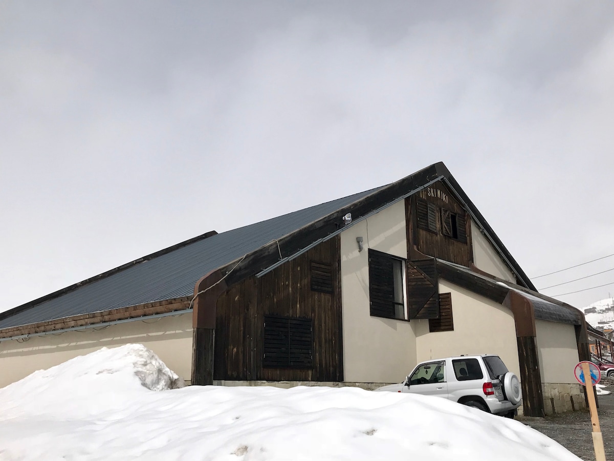 The exterior of a modern alpine house is depicted, showcasing a combination of dark wood and light-colored walls. The snow-covered ground surrounds the structure, highlighting its spacious roof and distinctive angled design. A parked vehicle is visible in front, adding context to the setting.