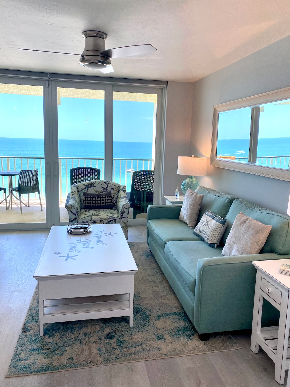 A bright living area is showcased, featuring a comfortable aqua sofa with decorative pillows and a patterned chair. A coffee table rests on a soft area rug. Sliding glass doors reveal expansive views of the beach and ocean beyond, allowing abundant natural light to fill the space.