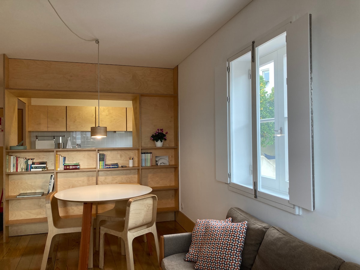 A light-filled living area features a round dining table surrounded by wooden chairs. A cozy sofa is positioned against the wall, adorned with a patterned cushion. Natural wood shelving in the background displays books and decorative items, while a pendant light hangs above the table.