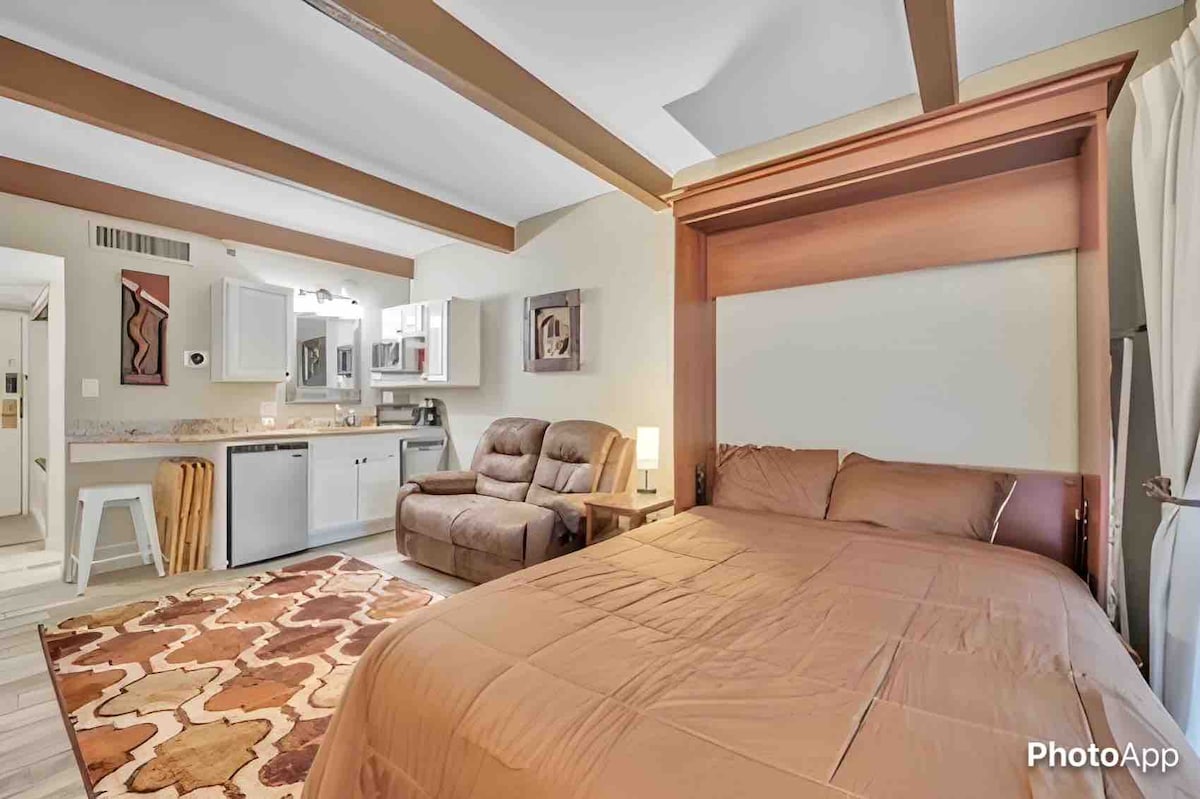 A studio space features a spacious bed with a warm-colored coverlet. A comfortable reclining chair sits adjacent, with a small lamp providing soft lighting. A compact kitchenette with modern appliances is visible in the background, enhancing the convenience of the area.