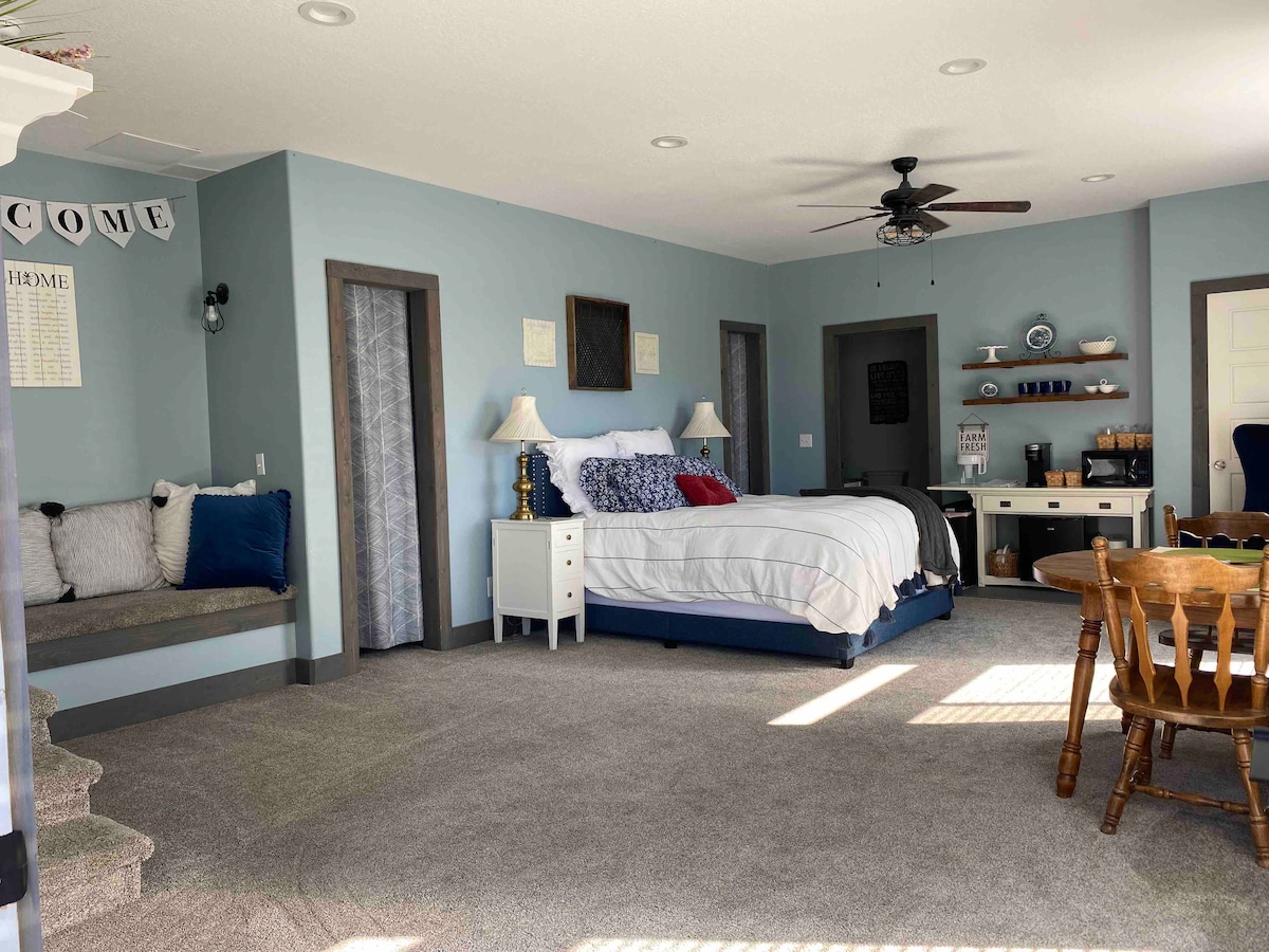 A spacious suite is shown, featuring a large bed with crisp white linens and accent pillows. A cozy reading nook with a bench sits against the wall. A dining area with wooden chairs and a desk with a coffee setup is visible. Soft light illuminates the room.