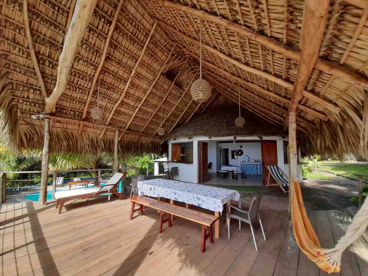 A spacious outdoor living area features a thatched roof made of palm leaves, providing natural shade. Wooden tables are arranged on the deck, accompanied by chairs and a hammock, creating a relaxed atmosphere. The kitchenette is visible in the background, with views of the pool area.