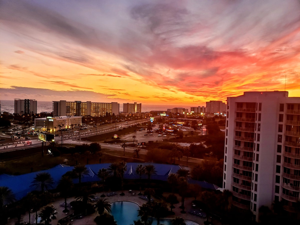 A stunning sunset illuminates the sky with vibrant hues of orange and pink. The view includes the coastline and buildings in the distance, while the resort pool and palm trees are visible below, contributing to a serene atmosphere.