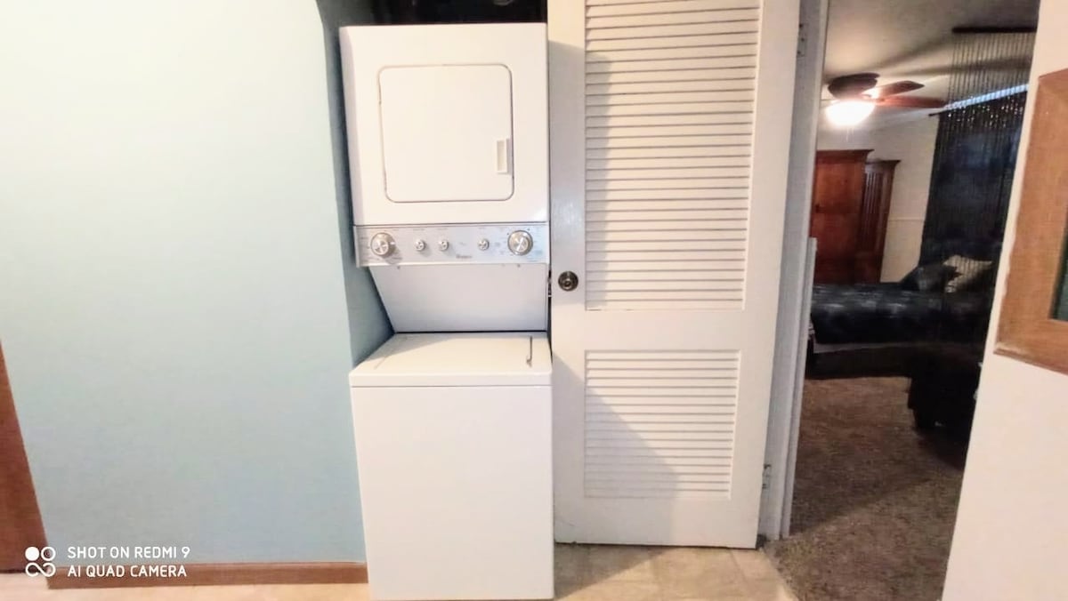 A stacked washer and dryer unit is positioned against a light blue wall, offering convenience within the apartment. A doorway with louvered doors is visible beside the unit, leading to a separate space. Soft, neutral flooring complements the overall design.