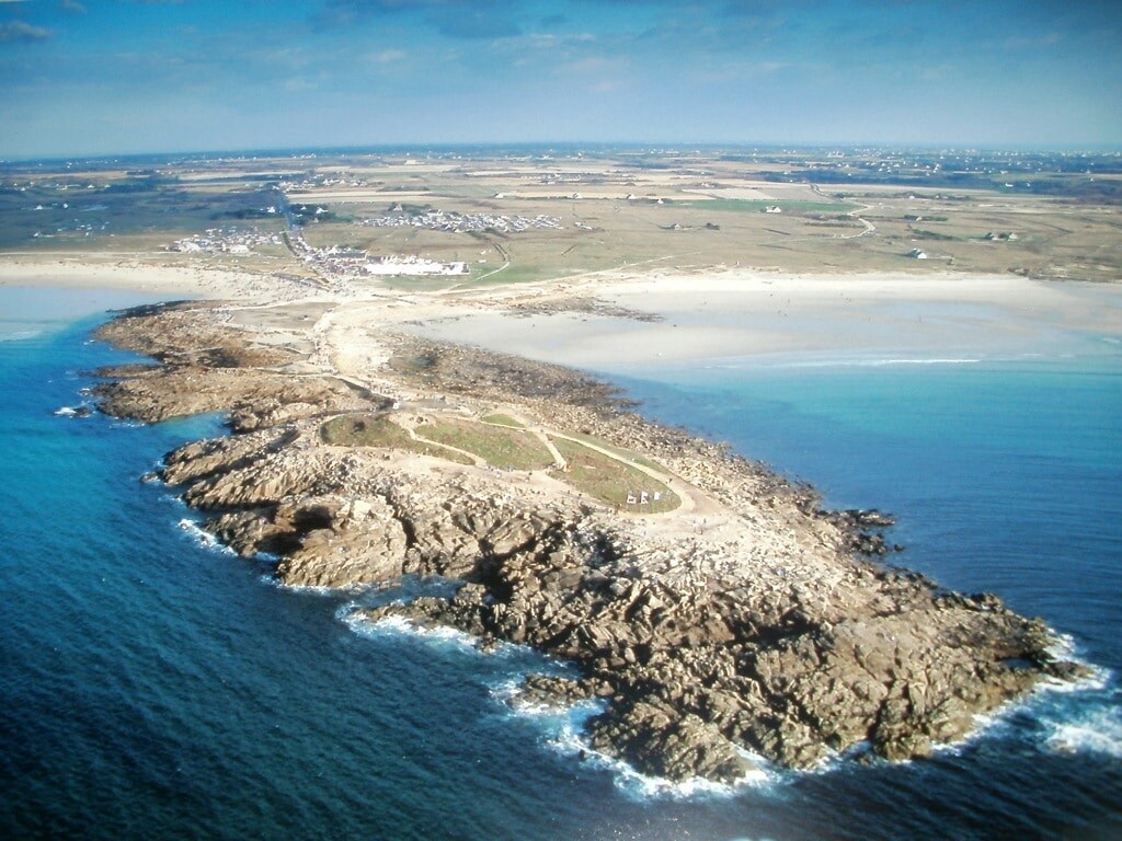 Aerial view of a rocky coastline extending into the ocean, surrounded by sandy beaches. The land features green patches and a pathway, with clear blue waters lapping against the shore. In the distance, the landscape transitions to flat fields and small settlements.