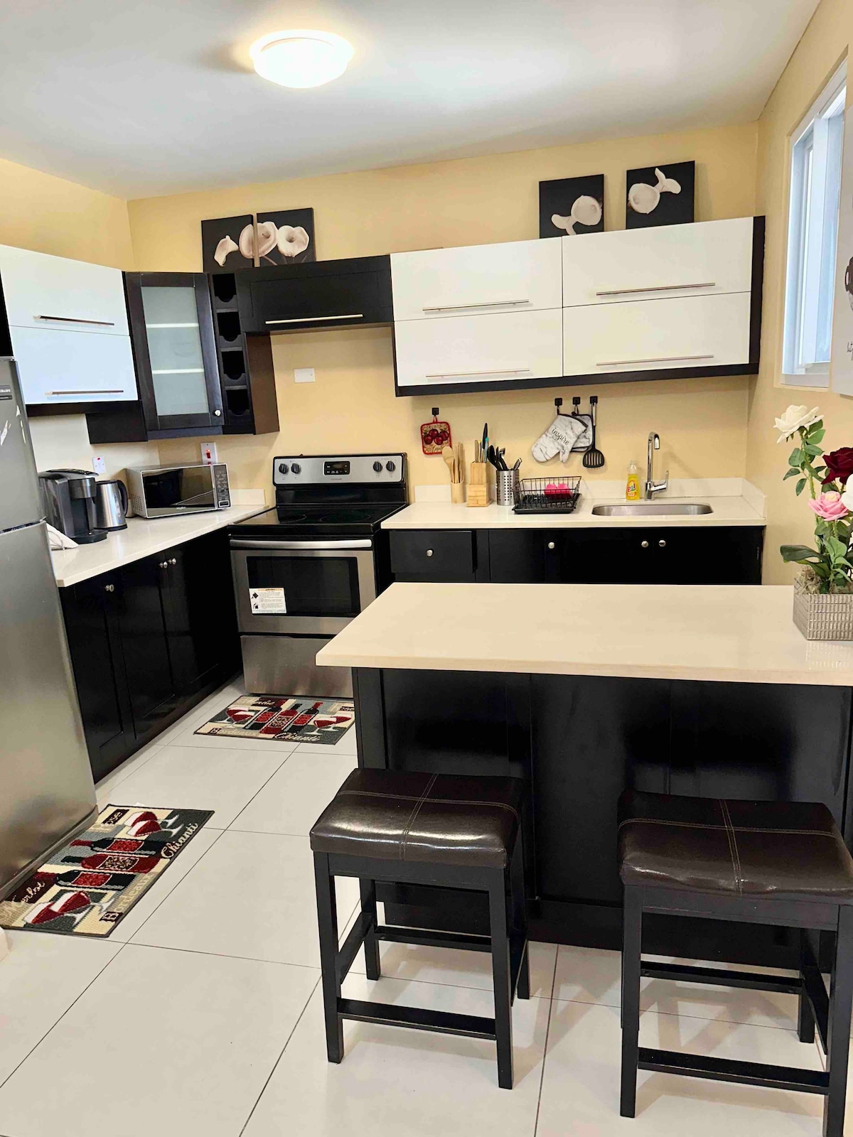 The kitchen features sleek black and white cabinetry, complemented by stainless steel appliances. A small island with two bar stools is present, with decorative accessories and a flower vase adding a touch of color. The countertops are clean and well-organized, providing an inviting cooking space.