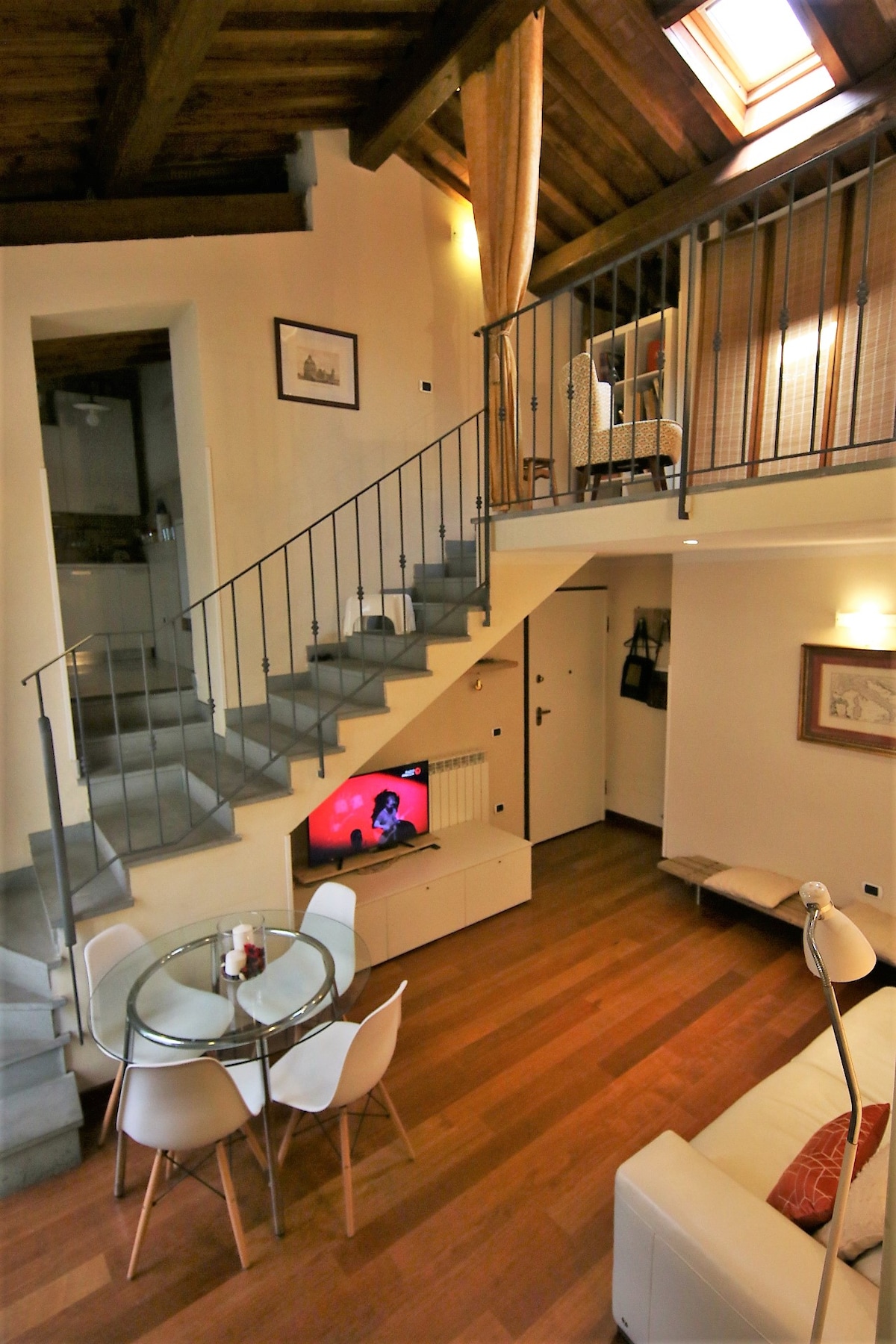 An open living area is highlighted by wooden beams reaching the ceiling, featuring a glass dining table surrounded by white chairs. A comfortable sofa faces a television, and a staircase leads to an upper level. Soft lighting creates a warm ambiance.
