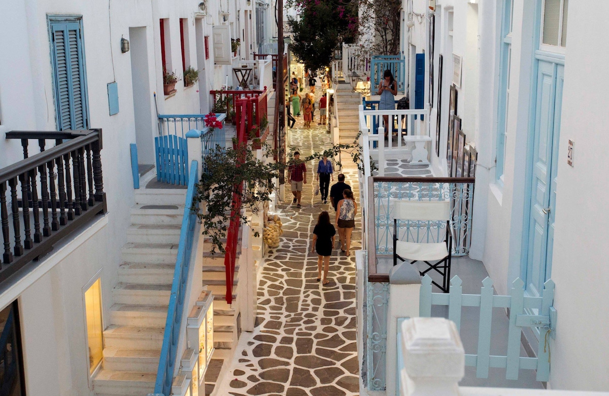 A vibrant pedestrian street in Mykonos is depicted, lined with charming white buildings. Blue accents enhance the traditional architecture. People stroll along the cobblestone path, with flowering plants visible on balconies. Soft lighting creates a welcoming atmosphere in the evening scene.