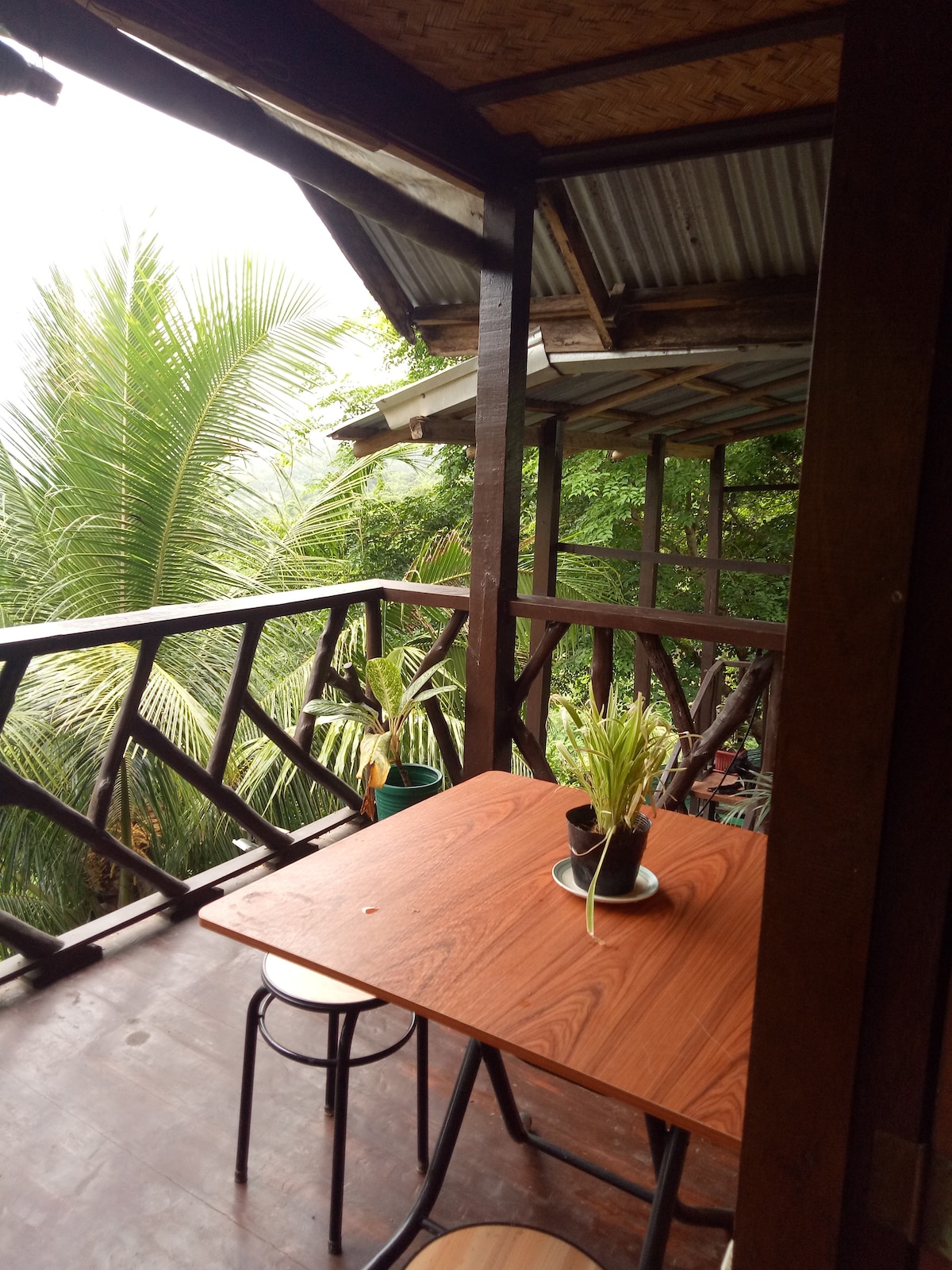 A wooden balcony features a sturdy table accompanied by two simple chairs. Lush greenery is visible in the background, enhancing the sense of being amidst nature. The structured roof provides shade, creating a comfortable area for relaxation.