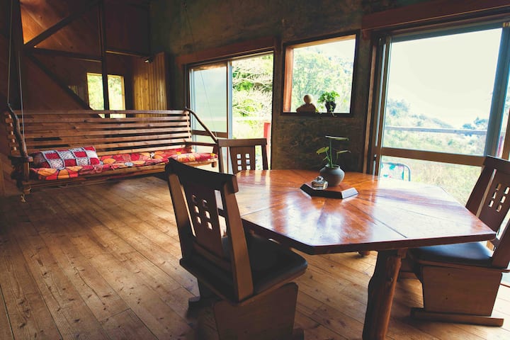 ห้องนั่งเล่นที่หันหน้าไปทางทิศใต้มีเก้าอี้ 6 ตัวที่โต๊ะนั่งเล่นเก้าอี้ 2 ตัวที่โต๊ะรับประทานอาหารและโซฟาที่นอนชิงช้าในร่มที่ทำด้วยมือของเจ้าของสะดวกสบายสำหรับผู้ใหญ่สูงสุด 2 คนห้องนั่งเล่นออกจากระเบียงพร้อมมหาสมุทรภูเขามหาสมุทรและภูเขา