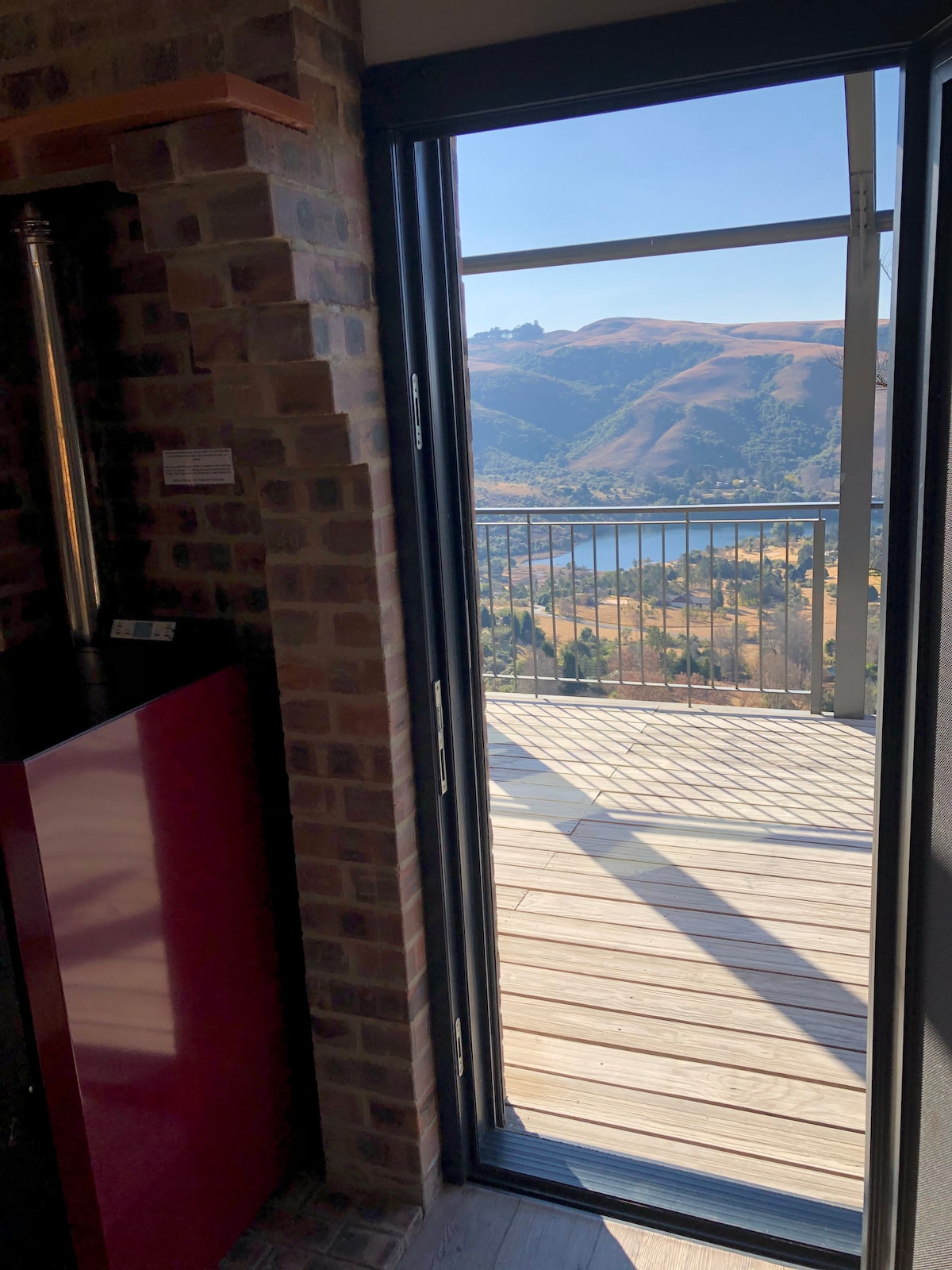 A doorway framed by a brick wall leads to a wooden deck with expansive views of rolling hills and a serene body of water. Natural light spills into the interior space, highlighting the contrasting textures of the brick and polished surfaces.
