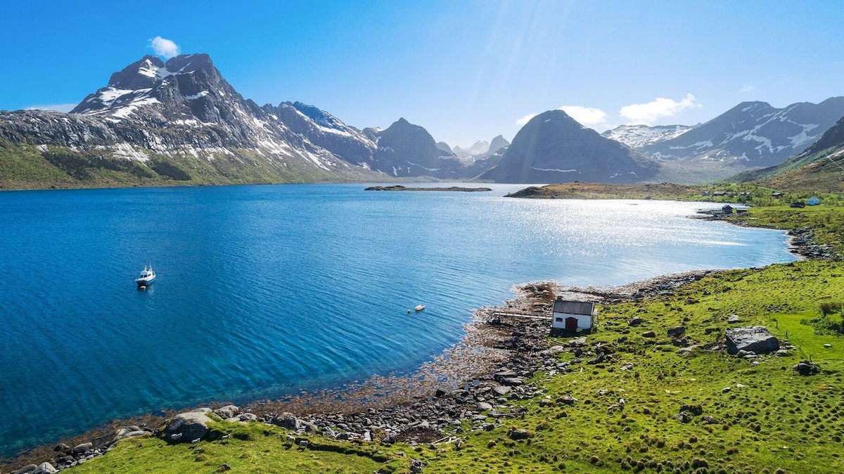 A scenic view of a calm blue lake surrounded by snow-capped mountains under a clear sky. A small boat is visible on the water, while a cozy building sits near the rocky shoreline, highlighting the tranquility of the natural landscape.