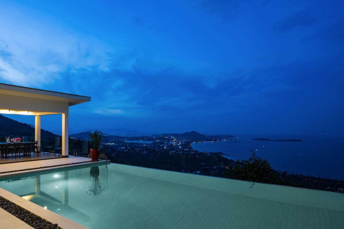 An infinity pool extends towards the horizon, reflecting the dusky blue sky. A spacious porch with dining furniture overlooks the water, with distant lights from the coastline creating a serene ambiance. Lush greenery frames the scene, enhancing the connection to nature.