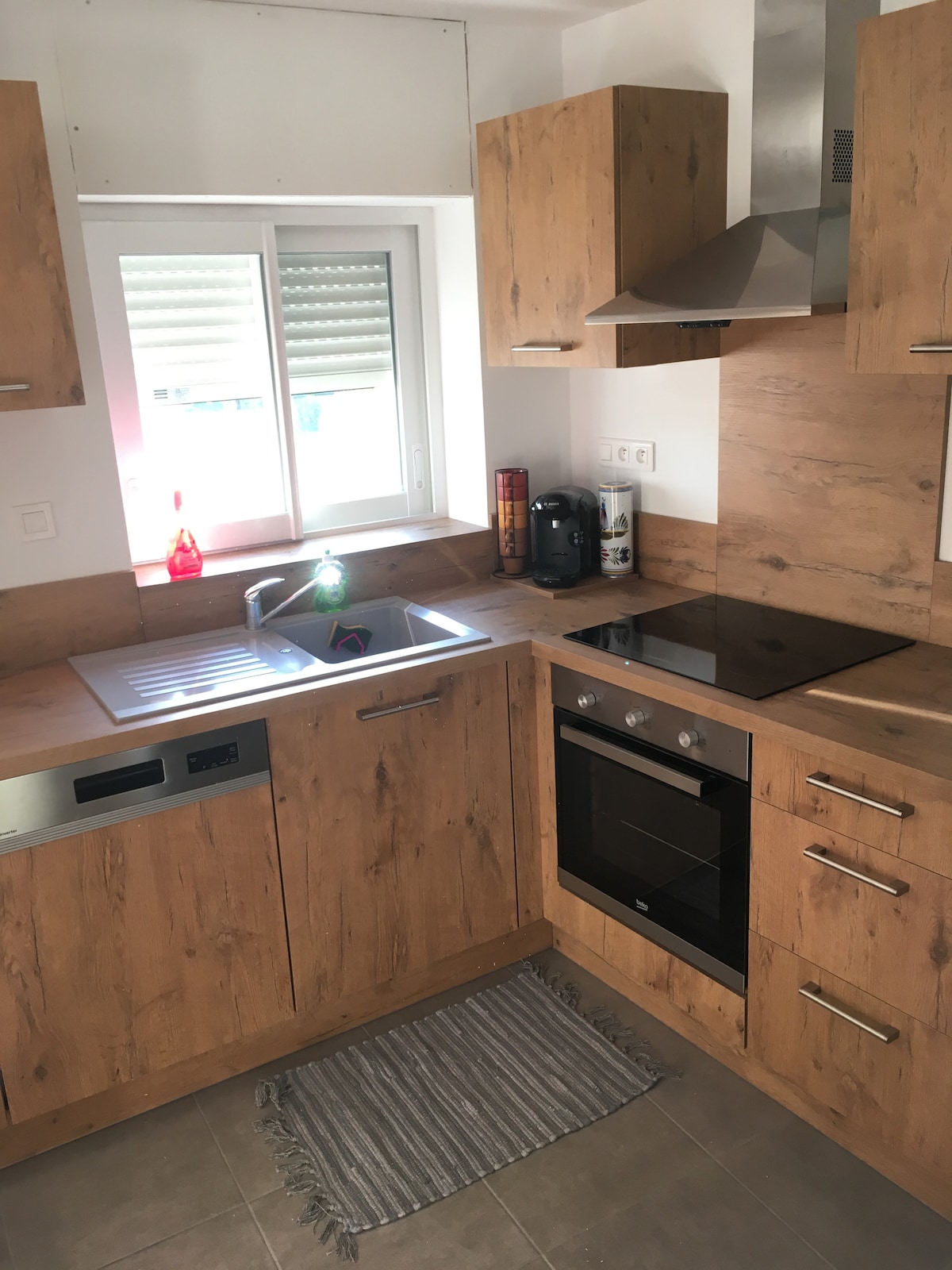 The kitchen features wood-patterned cabinetry, equipped with a sink, stovetop, oven, and coffee maker. A window allows natural light, while a small rug is placed on the tiled floor near the sink, enhancing the functional space.