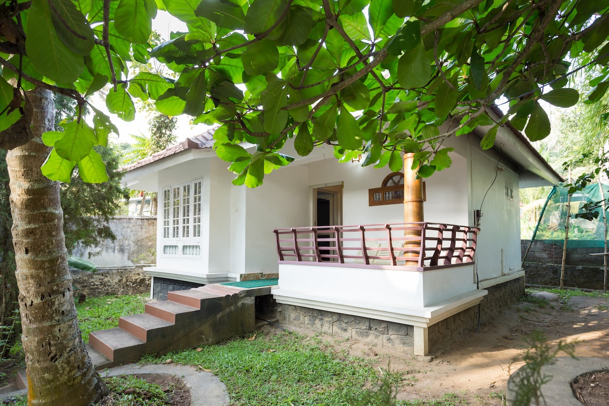 A charming villa is framed by lush green foliage, featuring a small porch with wooden railings. Steps lead up to the entrance, and large windows allow natural light to brighten the interior. The surrounding garden provides a serene outdoor space.