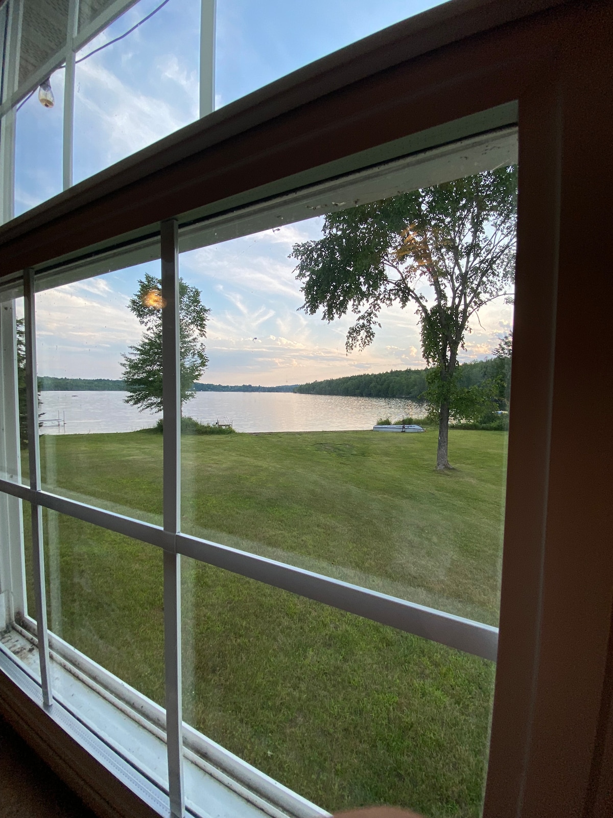 A scenic view is captured through a large window, revealing a calm lake surrounded by lush greenery. The sky showcases soft pastel hues, while the foreground features a well-maintained lawn extending to the water's edge.