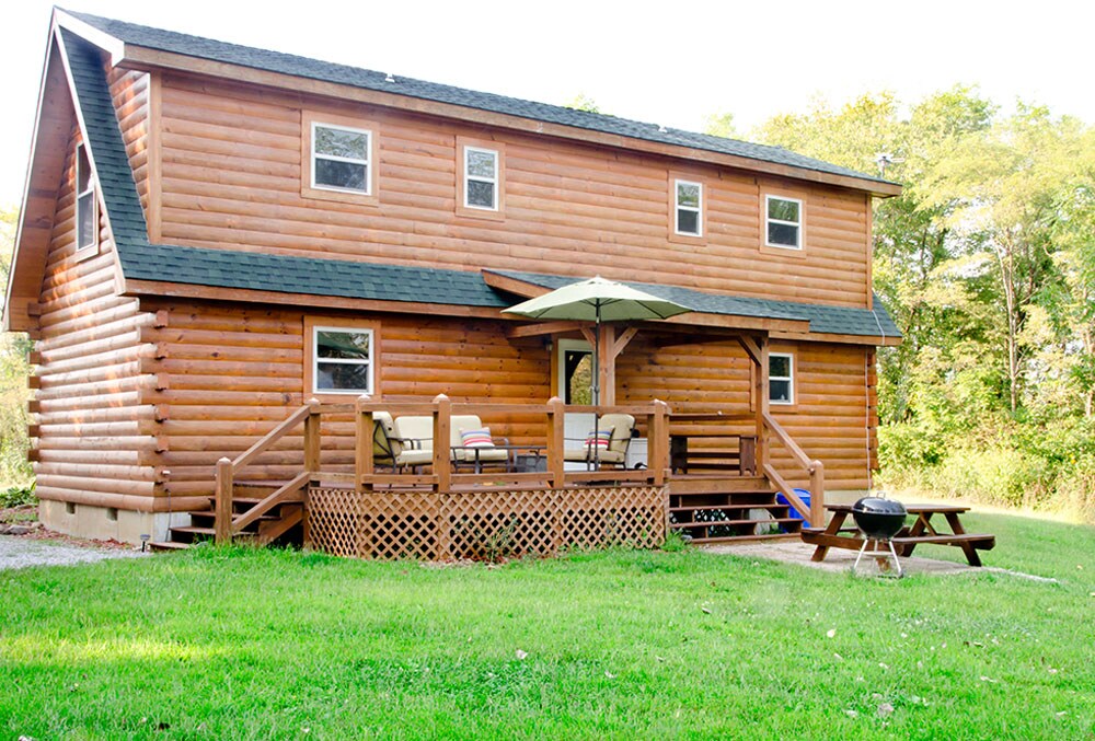 A rustic wooden lodge features a spacious front deck with outdoor seating under an umbrella. A barbecue grill is positioned nearby on a grassy area. The two-story structure is surrounded by trees, showcasing a serene natural setting.