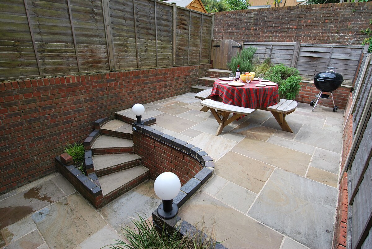 A small courtyard garden is visible, featuring a stone-paved surface with a set of stairs leading to a raised area. A circular table is set with dining essentials, surrounded by wooden benches. A charcoal barbecue is positioned nearby, with low garden lighting illuminating the space.