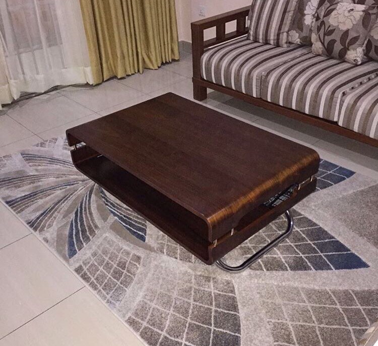 A low wooden coffee table with a sleek design sits on a patterned area rug. The table features a smooth surface and a lower shelf, while a striped sofa with floral cushions is positioned nearby. Drapes and curtains frame a window in the background.