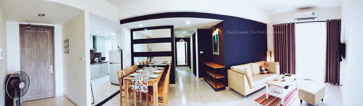 A spacious living and dining area features a table set for six, complemented by a cozy seating area with a sofa. The walls are painted in contrasting colors, and large windows allow natural light to fill the space. A fan and shelving unit are also visible.