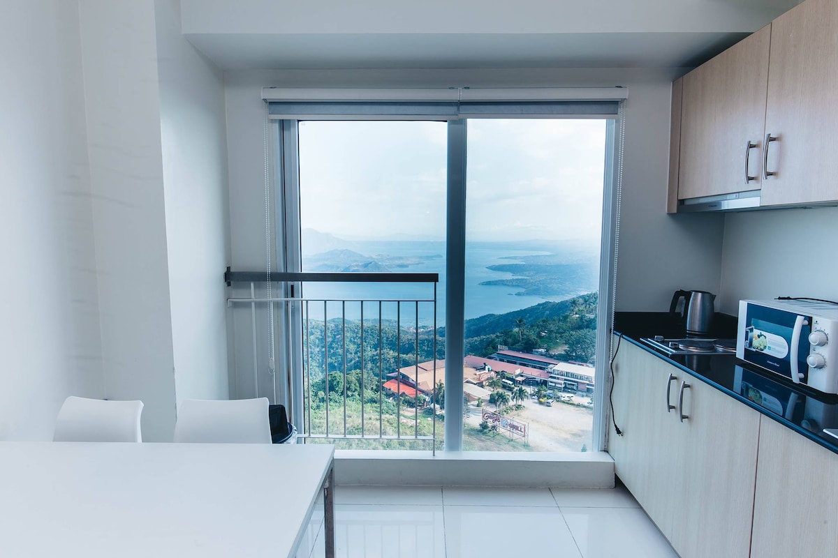 A modern kitchen area is visible, featuring a sleek countertop along with essential appliances including a kettle and microwave. A large window provides a panoramic view of Taal Lake and the surrounding landscape, filling the space with natural light.