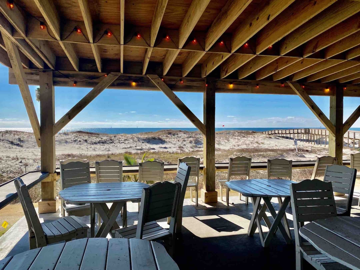 An open-air seating area features multiple white wooden tables under a wooden beam ceiling, with strings of lights enhancing the setting. A panoramic view of the sandy beach and ocean is visible through the railing, with beach access steps leading toward the water.