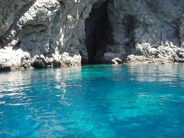 The image captures clear blue waters gently lapping against rocky shoreline, revealing a natural cave entrance nestled between the cliffs. Sunlight filters through the water, creating a calming interplay of light and shadows on the surface.
