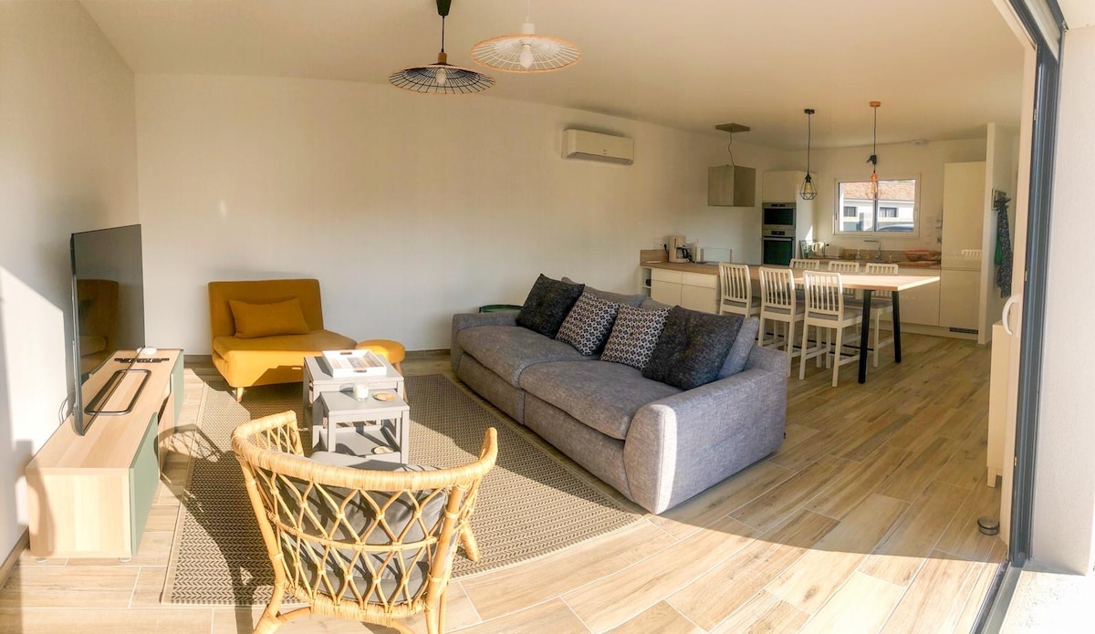 The living area is spacious, featuring a large grey sofa, a mustard yellow chair, and a flat-screen TV. A dining table with white chairs occupies the background, while wood and natural fibers enhance the warm ambiance. Windows provide ample natural light.