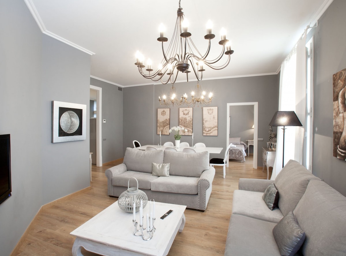 A spacious living area features a light grey sofa and a matching loveseat, accompanied by a chic coffee table. An elegant chandelier hangs from the ceiling, providing soft lighting. Artwork adorns the walls, and a dining area is visible in the background, complete with a table and chairs.