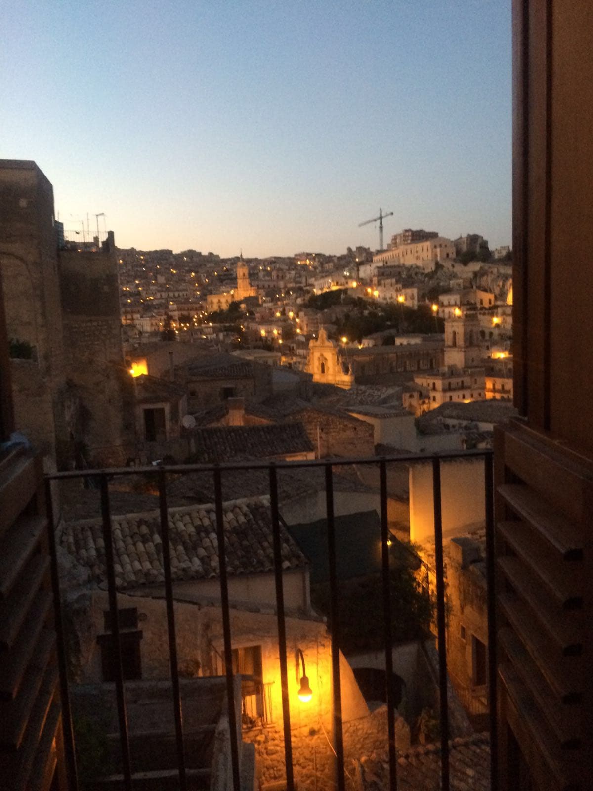 A view from the balcony features the illuminated cityscape of Modica at dusk. Soft lights illuminate the buildings, showcasing their historical architecture. A distant clock tower stands prominently among the rooftops, and a hint of the twilight sky enhances the serene atmosphere.