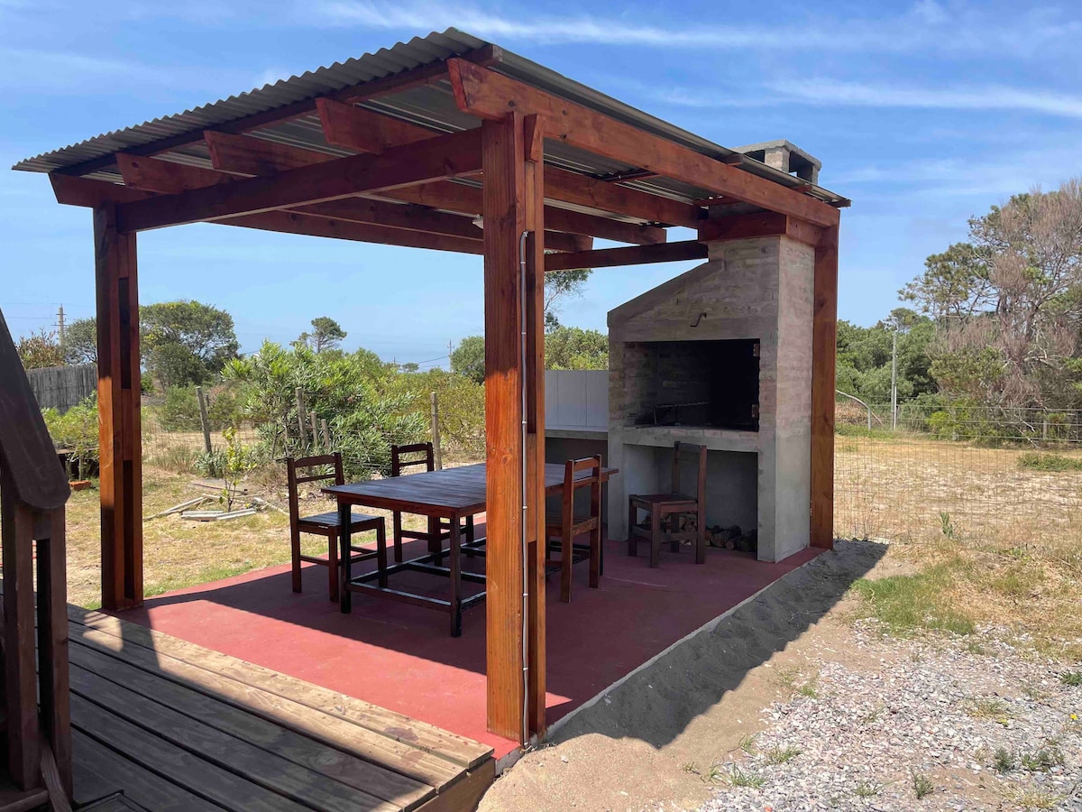 An outdoor cooking area features a sturdy wooden pergola with a barbecue grill and a stone fireplace. A wooden dining table with several chairs is positioned beneath the structure, offering space for gatherings. The surrounding landscape includes greenery and open space.